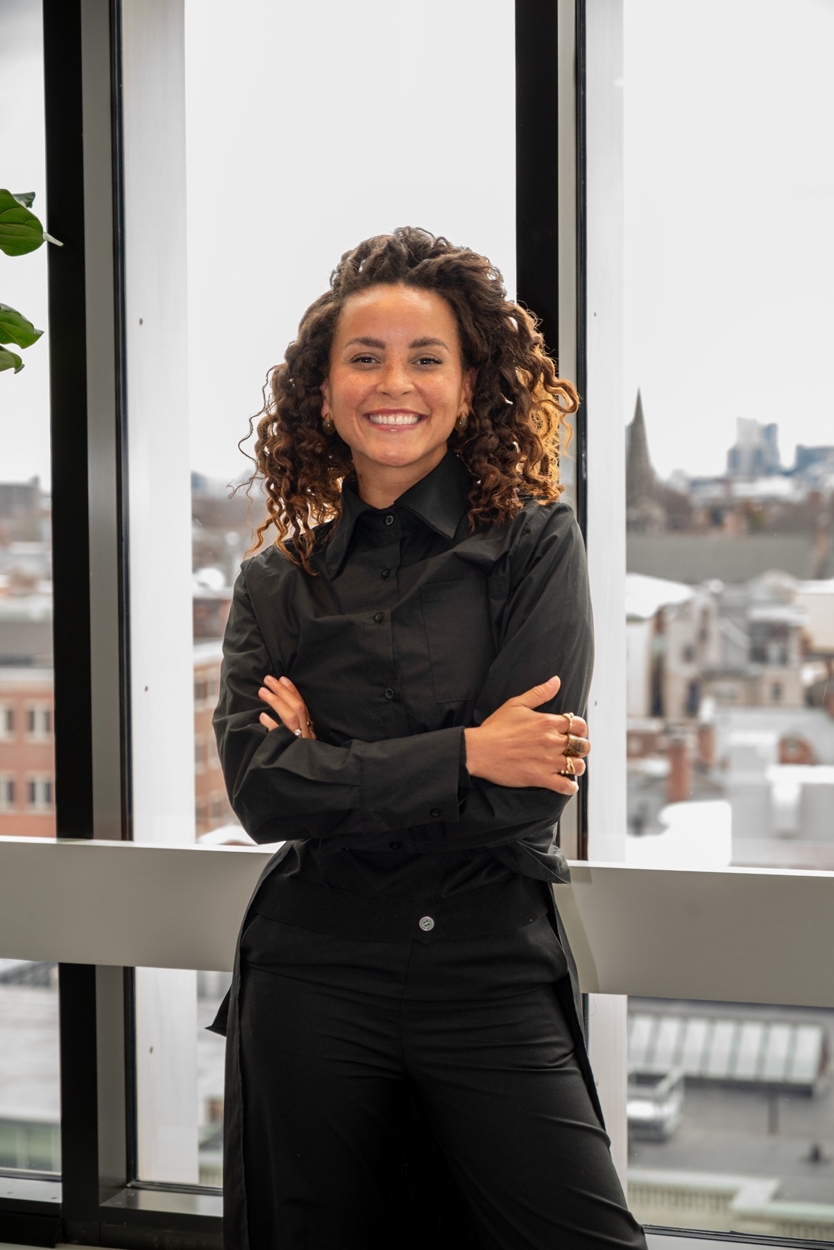 Domitila Barros sorrindo, de braços cruzados, diante de uma janela com vista da cidade